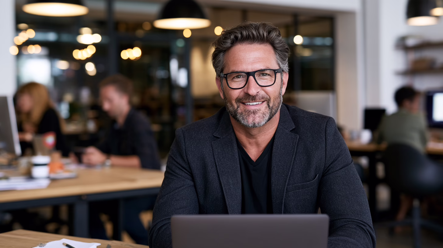 Smiling accounting firm manager working on a laptop in a busy, modern office with colleagues in the background.